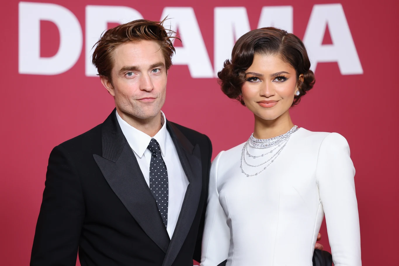 mężczyzna w czarnym smokingu i kobieta w eleganckiej białej sukni z kilkoma srebrnymi naszyjnikami stoją razem na tle czerwonej ściany z dużym białym napisem DRAMA