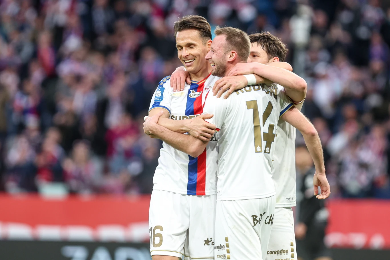 Trzech piłkarzy obejmuje się i cieszy po zdobyciu bramki podczas meczu na stadionie, na ich twarzach widać radość, a w tle rozmyci kibice dopingujący drużynę.