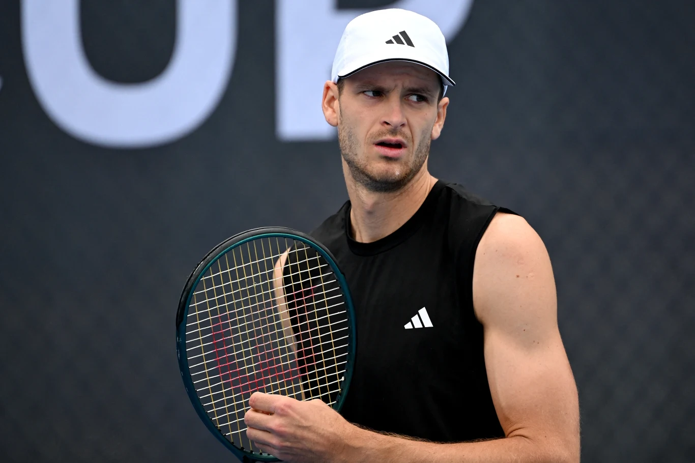 mężczyzna w sportowej czapce i czarnym bezrękawniku trzyma rakietę tenisową, skoncentrowany, stoi na tle ciemnego kortu tenisowego