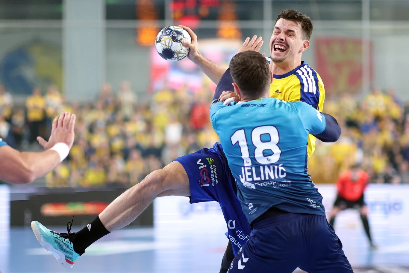 dynamiczna akcja podczas meczu piłki ręcznej, zawodnik w żółtej koszulce skacze z piłką nad broniącym go przeciwnikiem w niebieskiej koszulce, widoczne skupienie i emocje na twarzach graczy