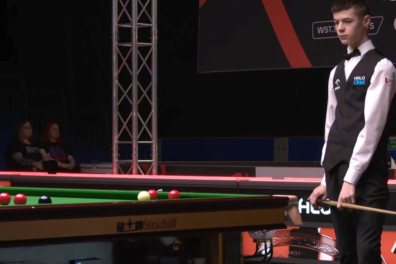 młody snookerzysta w kamizelce i muchą stoi przy stole bilardowym, trzymając kij i uważnie obserwuje rozmieszczenie bil na zielonym suknie podczas turnieju