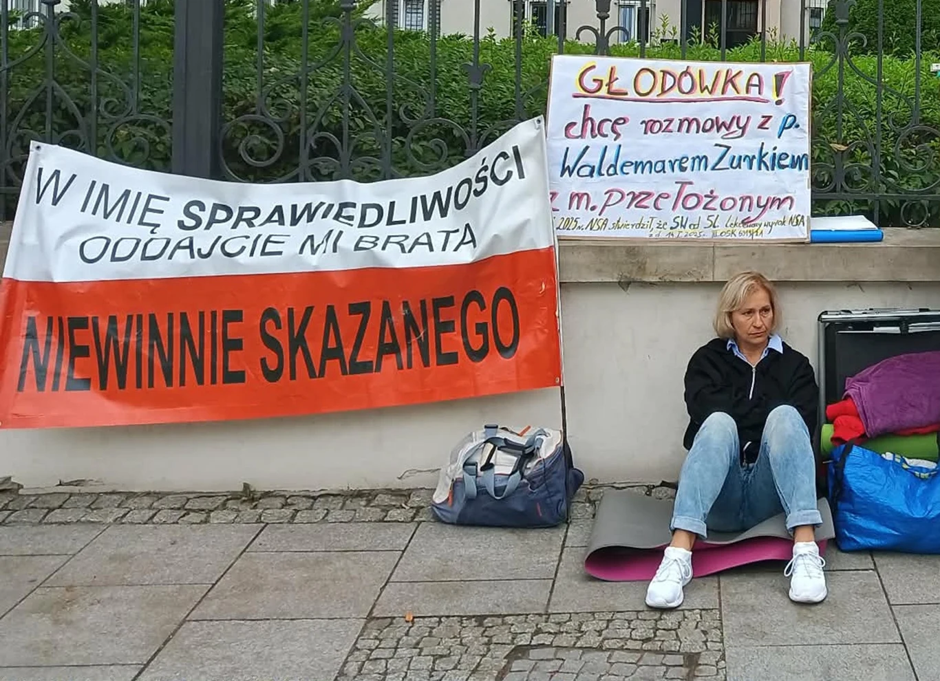 kobieta siedząca na chodniku obok rozłożonych rzeczy, przed barierkami z wywieszonymi transparentami domagającymi się sprawiedliwości i uwolnienia niewinnie skazanego, z informacją o głodówce i żądaniu rozmowy z przedstawicielem