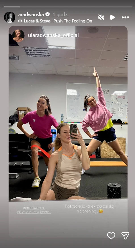 Trzy kobiety w kolorowych strojach sportowych pozują uśmiechnięte na tle sali treningowej, z matami do ćwiczeń i sprzętem fitness w tle; jedna z nich siedzi na macie, dwie stoją obok i wykonują dynamiczne gesty.