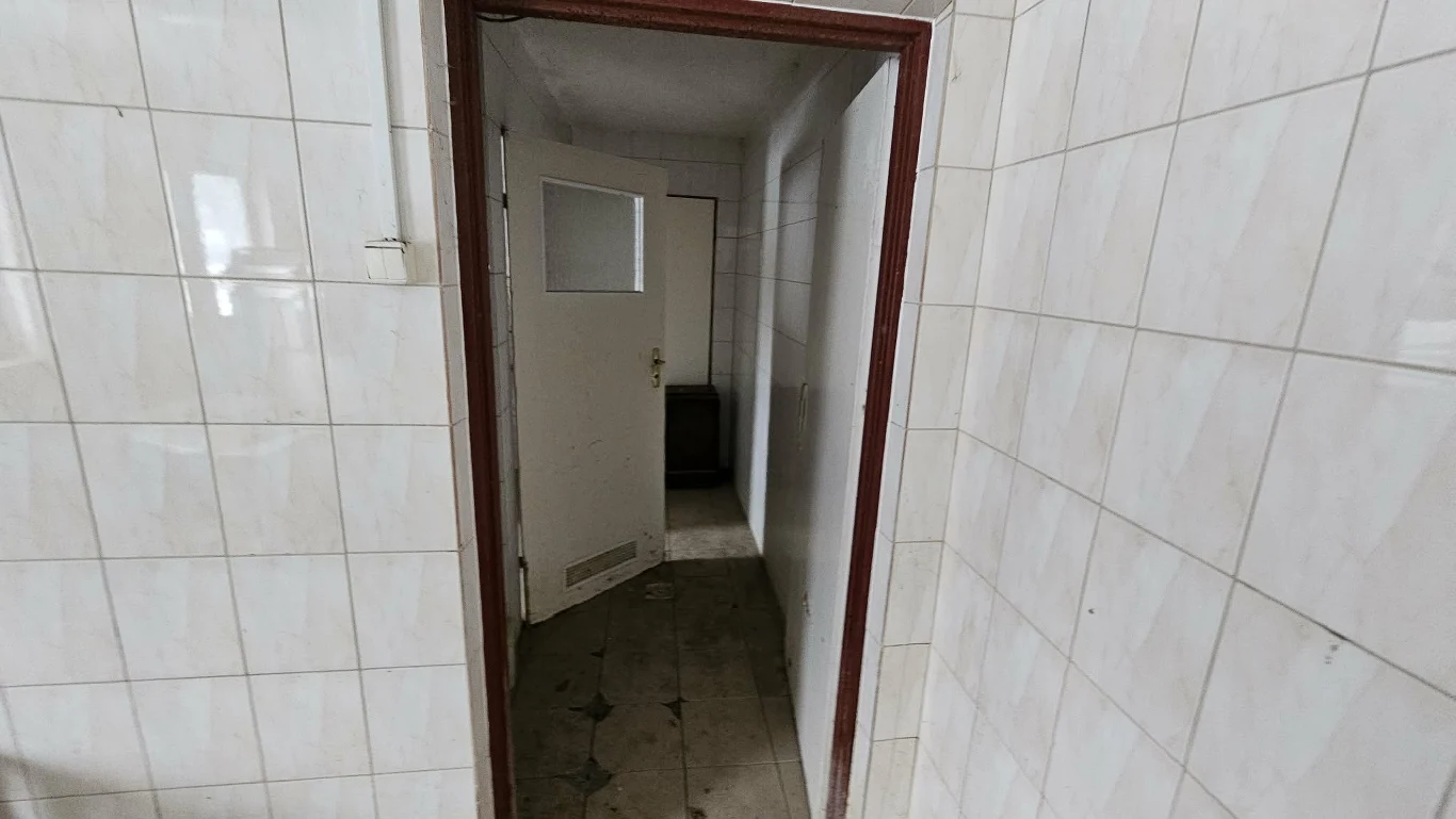 wejście do małego, zaniedbanego pomieszczenia z białymi płytkami na ścianach i uchylonymi drzwiami, prowadzącymi do kolejnego ciemnego pokoju