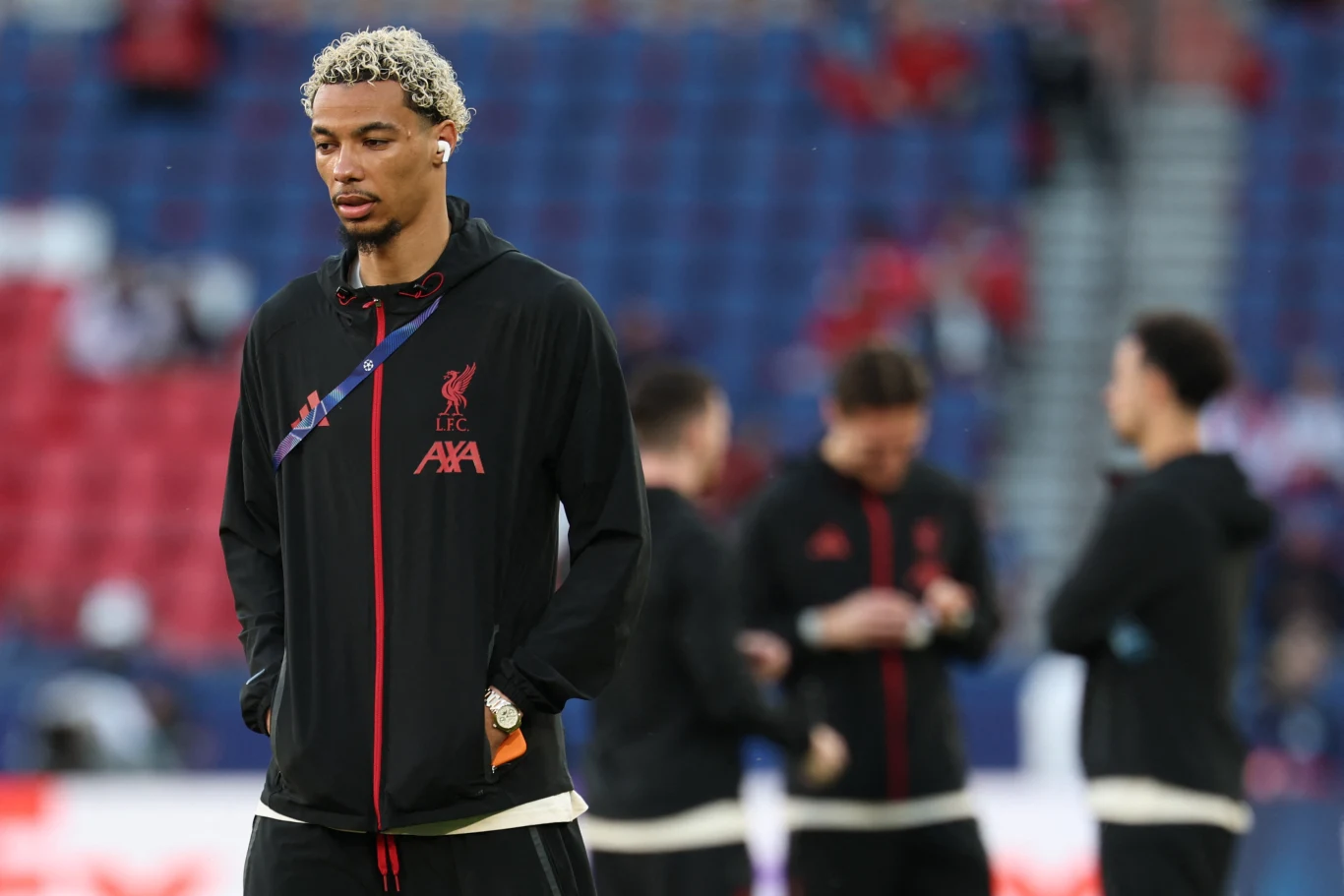 młody mężczyzna w dresie Liverpool FC z charakterystycznym jasnym afro spaceruje po murawie stadionu, w tle kilku innych piłkarzy rozmawia ze sobą lub korzysta ze smartfonów