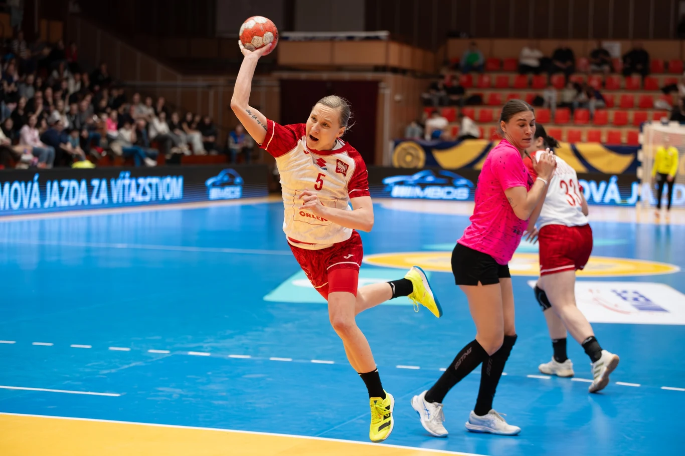 zawodniczka w białoczerwonym stroju piłki ręcznej wykonuje dynamiczny rzut w kierunku bramki, podczas gdy obrończyni w różowej koszulce próbuje ją zablokować na boisku sportowym