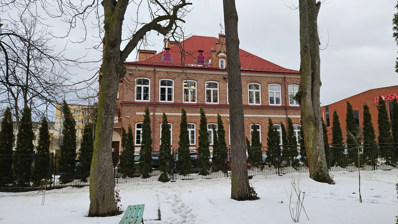 Ceglany budynek o dwóch kondygnacjach z czerwonym dachem, otoczony rzędem wysokich iglaków i kilkoma dużymi drzewami na zaśnieżonym terenie parkowym, z zieloną ławką ustawioną przed budynkiem.