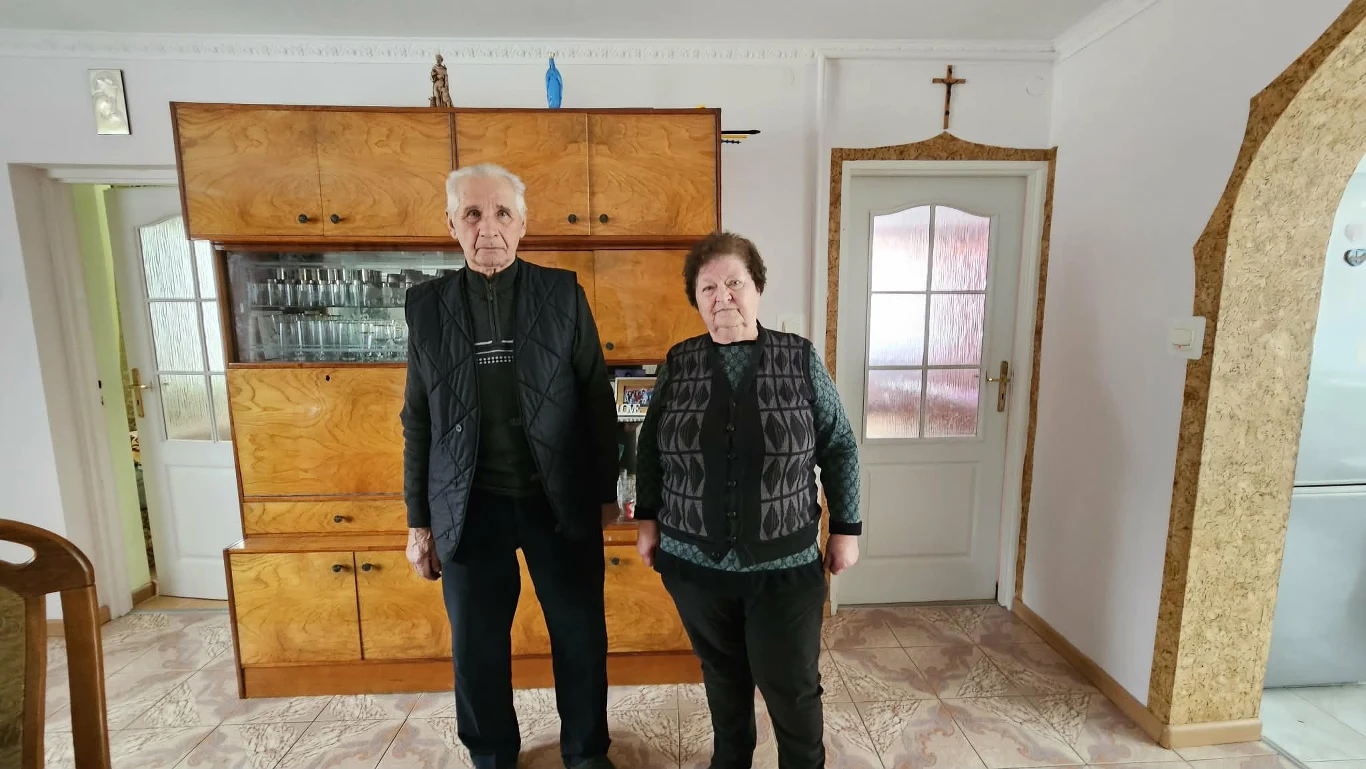 starsza kobieta i mężczyzna stoją obok siebie w jasnym, schludnym pokoju z drewnianą meblościanką na tle, na której stoją dekoracje i krzyż wiszący nad drzwiami