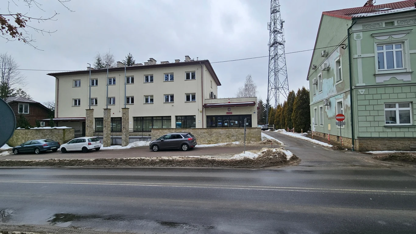 trzypiętrowy, jasny budynek z przeszklonym parterem i kilkoma zaparkowanymi samochodami przed wejściem, z prawej strony widoczny zielony budynek oraz metalowa wieża telekomunikacyjna w tle; na chodniku i krawężniku leży niewielka ilość śniegu