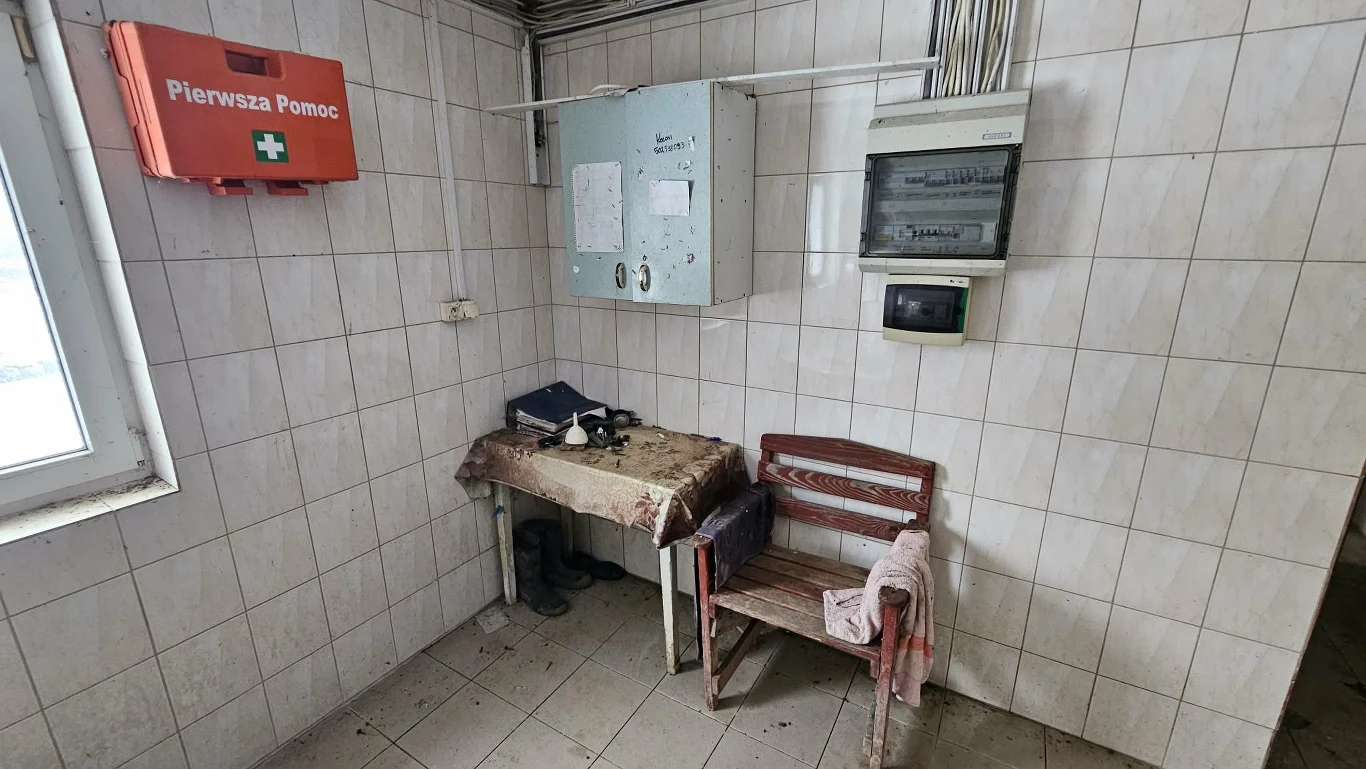 Zaniedbane pomieszczenie z kafelkowanymi ścianami, zniszczonym stołem, metalowym krzesłem z narzuconą szmatką, skrzynką pierwszej pomocy na ścianie oraz skrzynkami elektrycznymi i licznikiem zamontowanymi nad stołem.