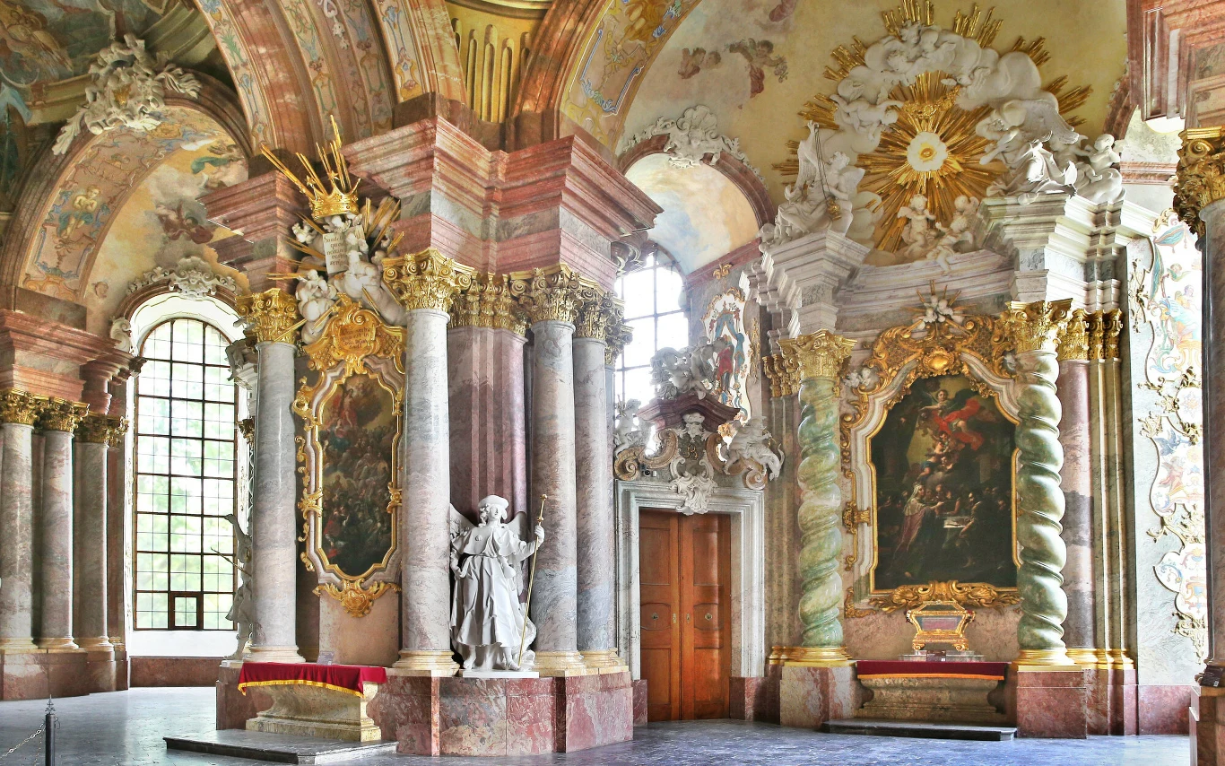 Barokowe wnętrze kościoła z bogato zdobionymi kolumnami, złoceniami, dużymi obrazami religijnymi oraz białą rzeźbą postaci świętej na tle marmurowych detali i bogato ornamentowanego sufitu.