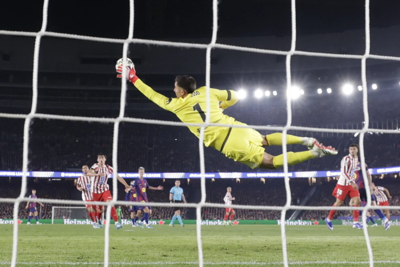 bramkarz w żółtym stroju rzuca się w powietrzu z wyciągniętą ręką broniąc piłkę lecącą w kierunku bramki, w tle kilku zawodników drużyn w pasiastych i jednolitych strojach oraz sędzia na środku boiska, silne oświetlenie stadionu