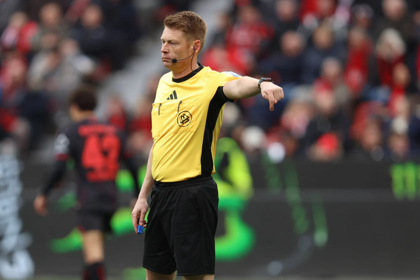 sędzia piłkarski w żółtej koszulce i czarnych spodenkach wskazuje kierunek gry, trzymając w ręku gwizdek, na tle nieostrej publiczności i zawodników