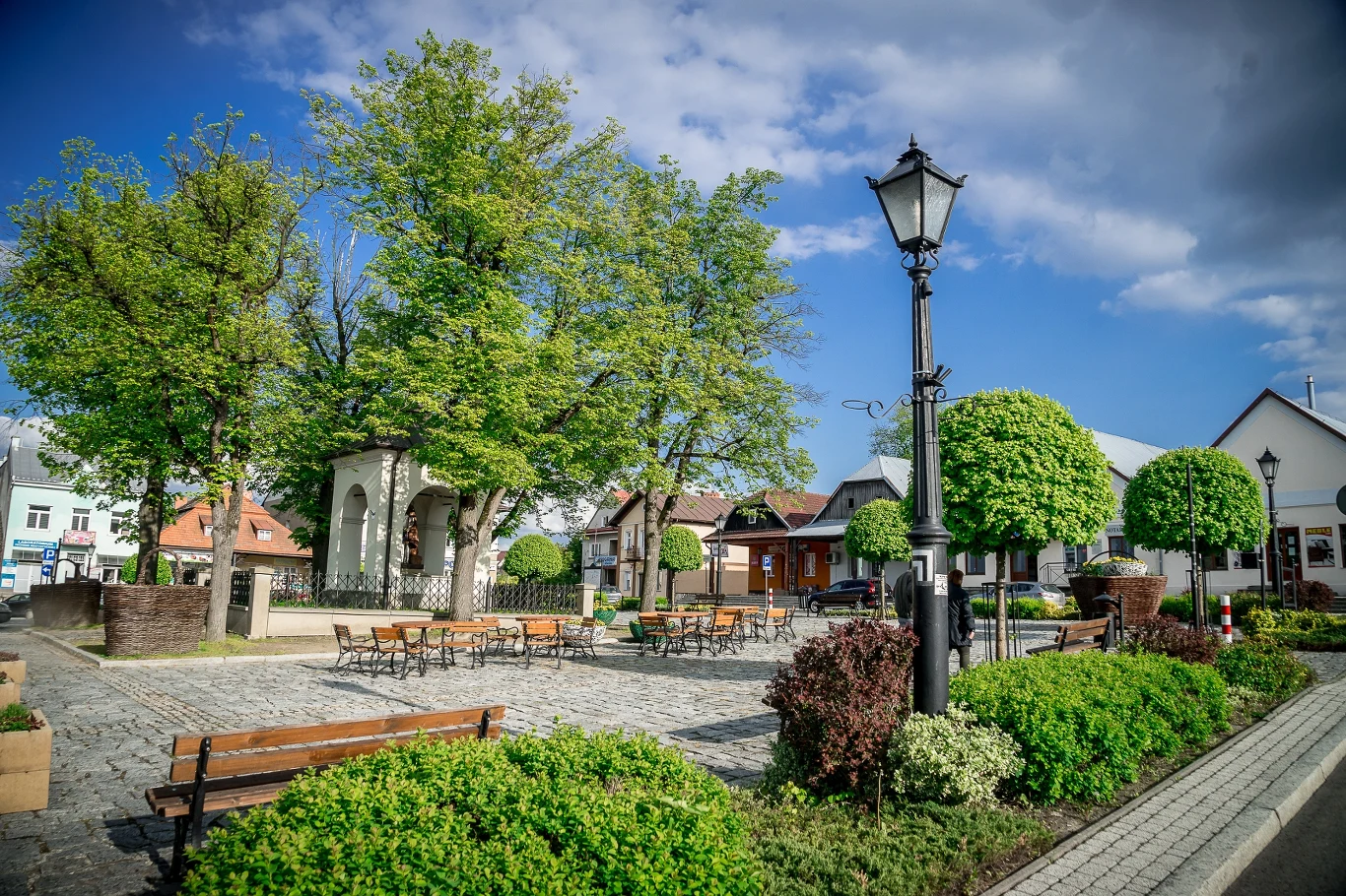 mały rynek miejski z brukowanym placem, ławkami, latarnią i zadbaną zielenią, w centrum kilka stolików z krzesłami pod dużymi drzewami, w tle niskie budynki mieszkalne i białe altany