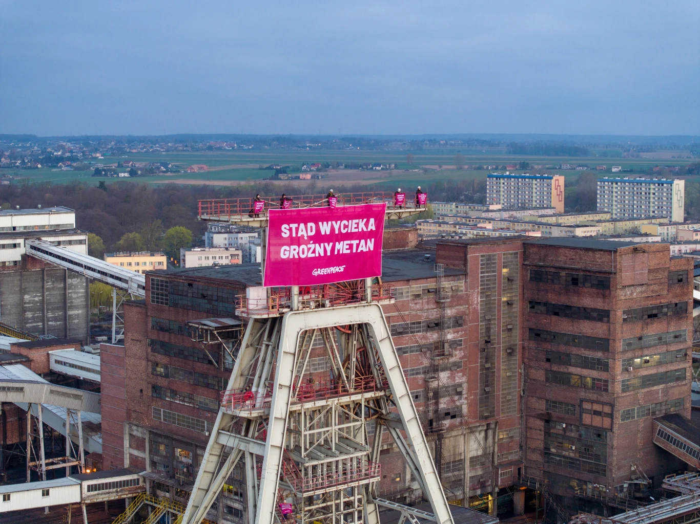 Kompleks przemysłowy kopalni z centralnie umieszczoną wieżą szybową, na której zamieszczony jest duży, różowy baner z napisem „STĄD WYCIEKA GROŹNY METAN”, na tle bloków mieszkalnych i rozległych pól w oddali.