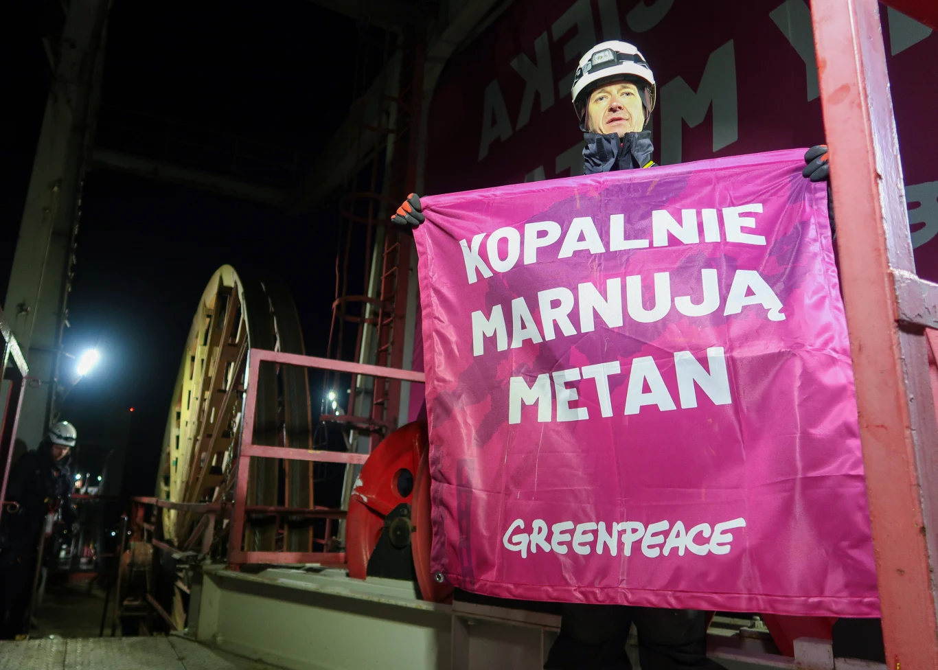 osoba w kasku ochronnym trzymająca różowy transparent z białym napisem 'Kopalnie marnują metan Greenpeace' na tle przemysłowego wyposażenia kopalni