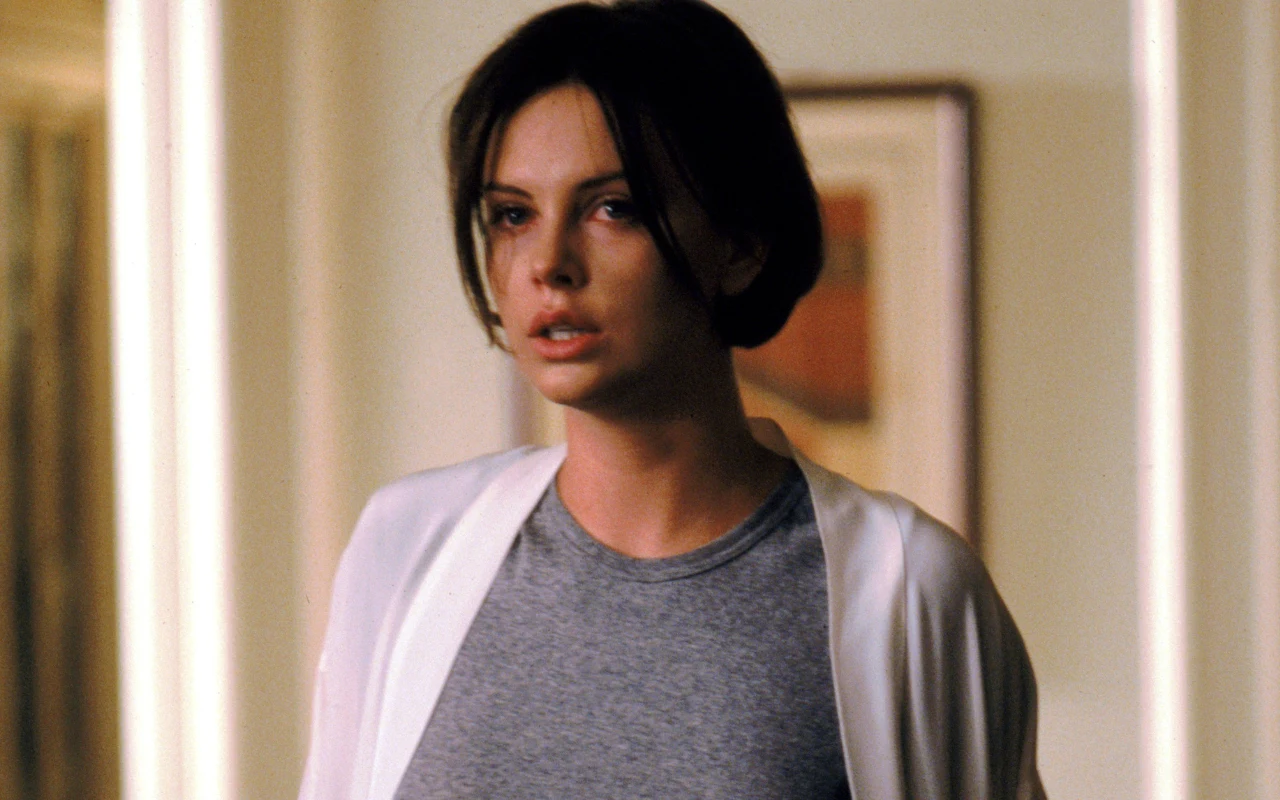 Charlize Theron w swojej przełomowej roli z 1997 roku w filmie "Adwokat diabła" Młoda kobieta o krótkich, ciemnych włosach ubrana w szary t-shirt i białą narzutkę, stoi na tle jasnej ściany z zawieszonym, abstrakcyjnym obrazem w ramce.