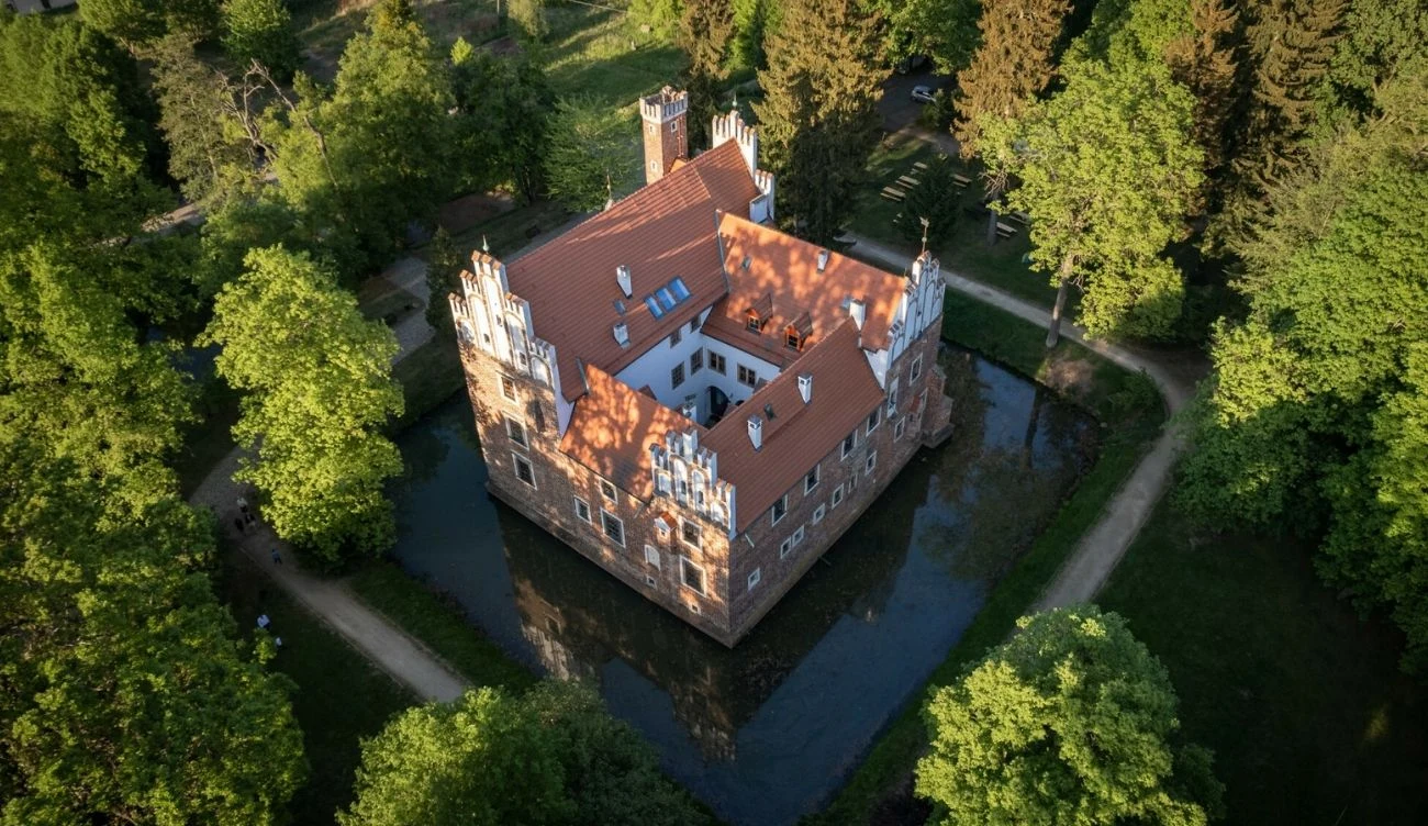 Zabytkowy zamek otoczony fosą i zwarty park z wysokimi drzewami, jasne mury z czerwonym dachem, wąskie wieżyczki w narożnikach oraz szerokie ścieżki prowadzące wokół budowli.