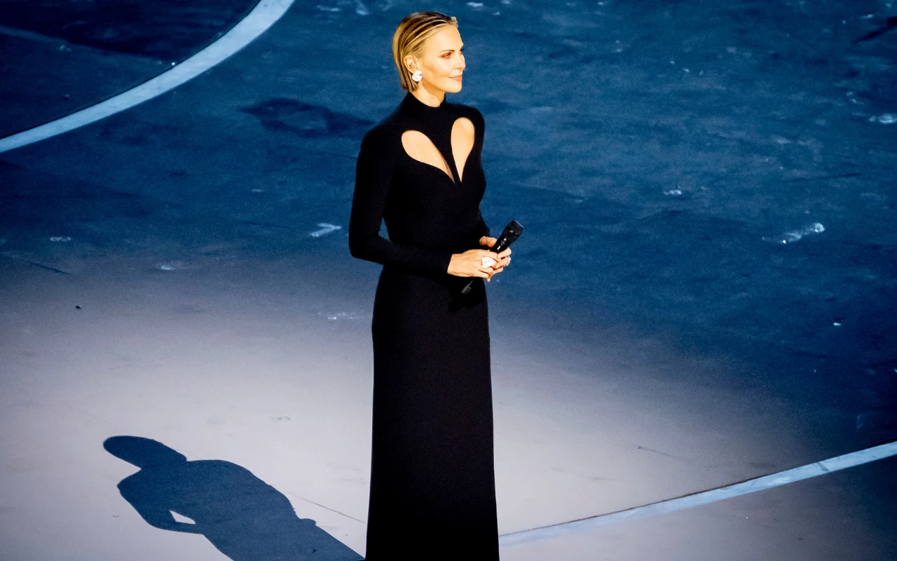 Charlize Theron na ceremonii otwarcia Zimowych Igrzysk Olimpijskich w 2026 roku kobieta w eleganckiej, długiej czarnej sukni z wycięciami na dekolcie, stojąca na niebiesko oświetlonej scenie i trzymająca mikrofon