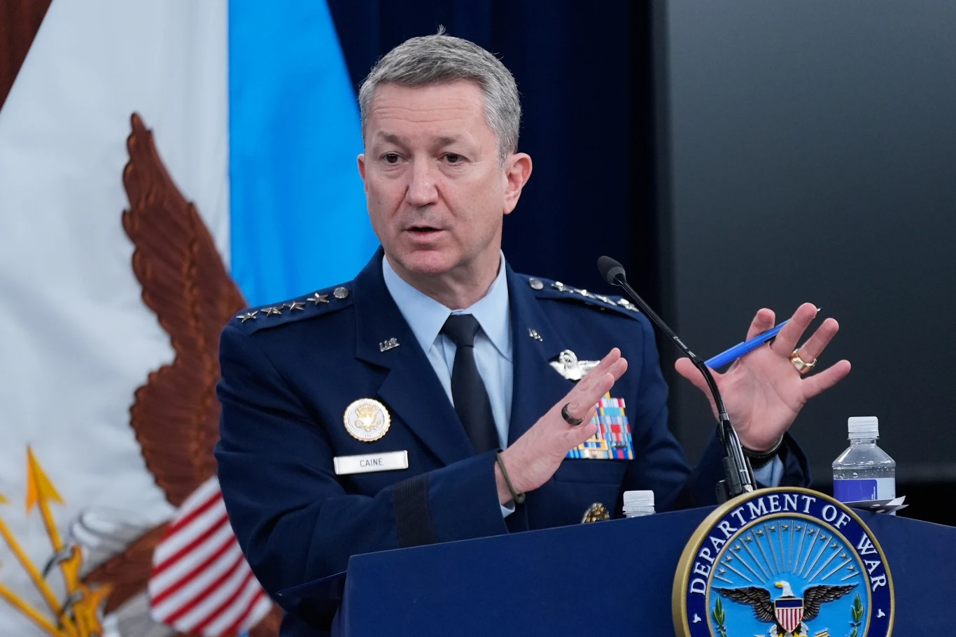 Wysoki rangą wojskowy w granatowym mundurze ozdobionym medalami i odznaczeniami stoi za mównicą z napisem 'Department of War', gestykulując rękami podczas przemówienia; w tle widoczne flagi państwowe i emblematy wojskowe.