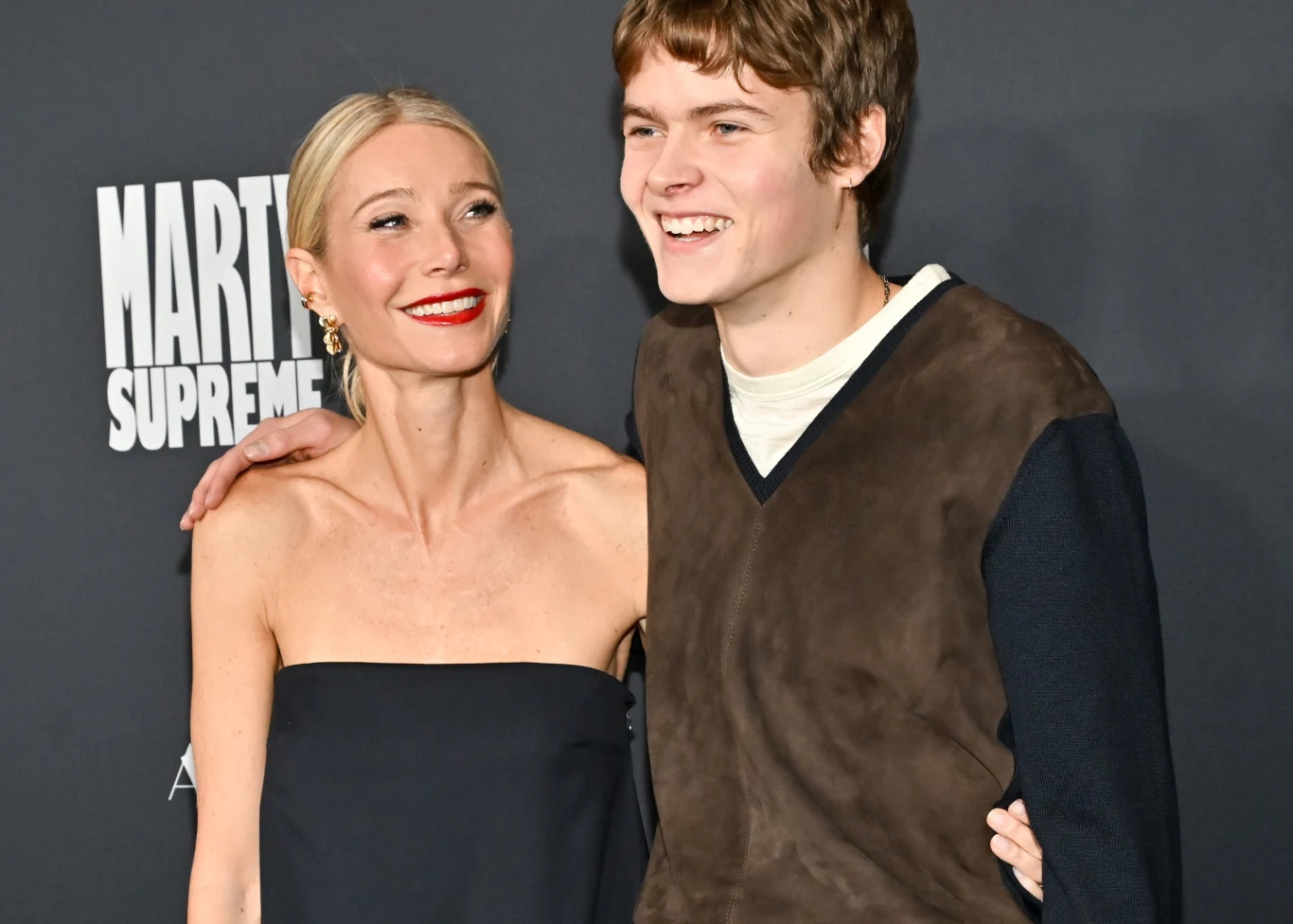 Kobieta w czarnej, eleganckiej sukni z czerwonymi ustami i blond włosami upiętymi do tyłu stoi obok uśmiechniętego młodego mężczyzny w brązowym bezrękawniku nałożonym na czarną bluzę; oboje pozują razem na tle ciemnej ścianki z jasnym logo tekstowym.