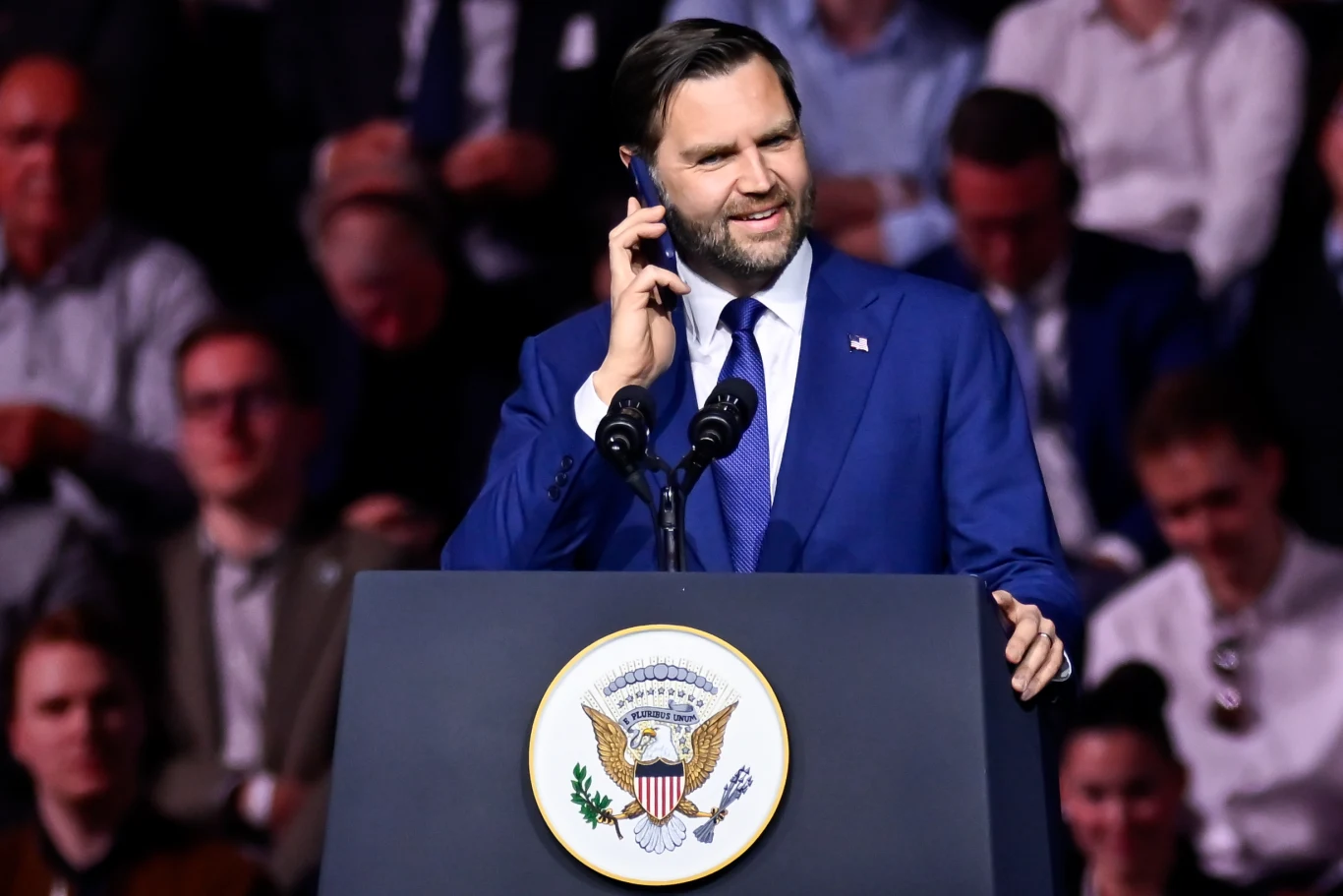 Mężczyzna w garniturze i pod krawatem stoi za mównicą z herbem prezydenta Stanów Zjednoczonych, uśmiecha się i rozmawia przez telefon, wokół liczna publiczność w półcieniu.