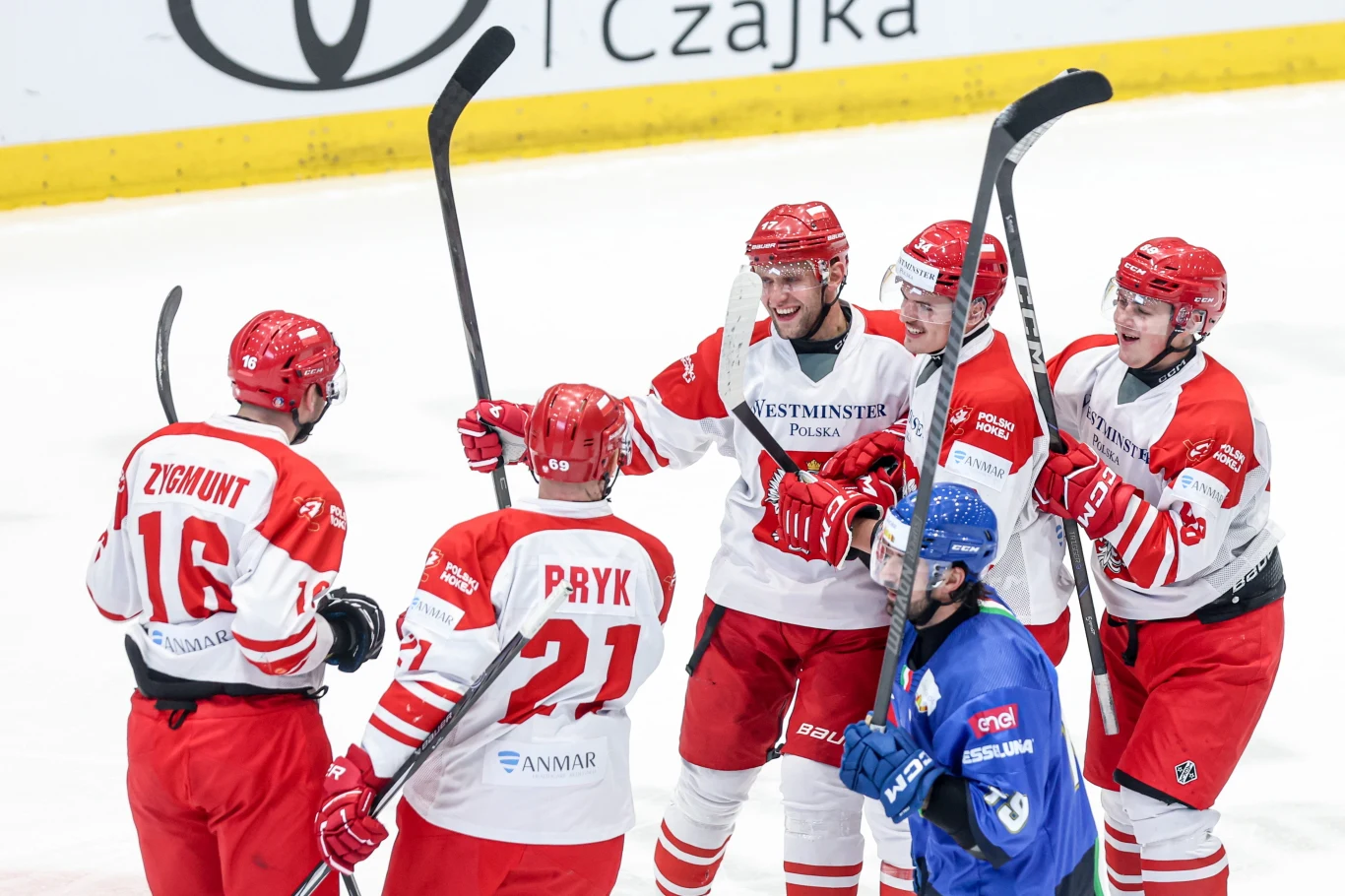 Reprezentacja Polski w hokeju wystąpi w MŚ 2026 Dywizji IA przed publicznością w Sosnowcu hokeiści w biało-czerwonych strojach zgromadzeni razem, świętują zdobycie bramki, obok nich zawodnik w niebieskiej koszulce z opuszczoną głową
