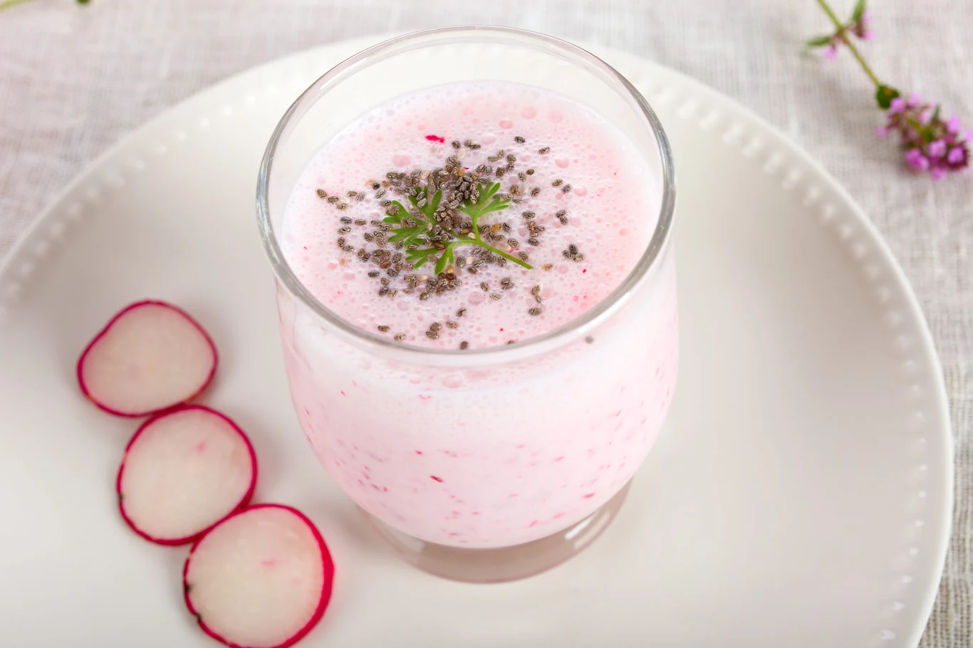 Różowy koktajl rzodkiewkowy na kefirze Szklanka różowego koktajlu z rzodkiewki i kefiru, posypana chia i natką, obok plasterki rzodkiewki na talerzu.