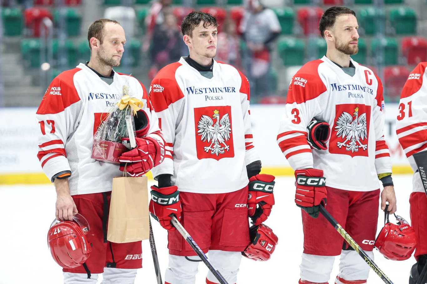 Trzech hokeistów w biało-czerwonych strojach reprezentacji Polski stoi obok siebie na lodowisku, każdy trzyma kij hokejowy i czerwony kask, jeden z zawodników dodatkowo trzyma torbę oraz nagrodę, tło stanowią pustawce trybuny z zielono-czerwonymi krzes...