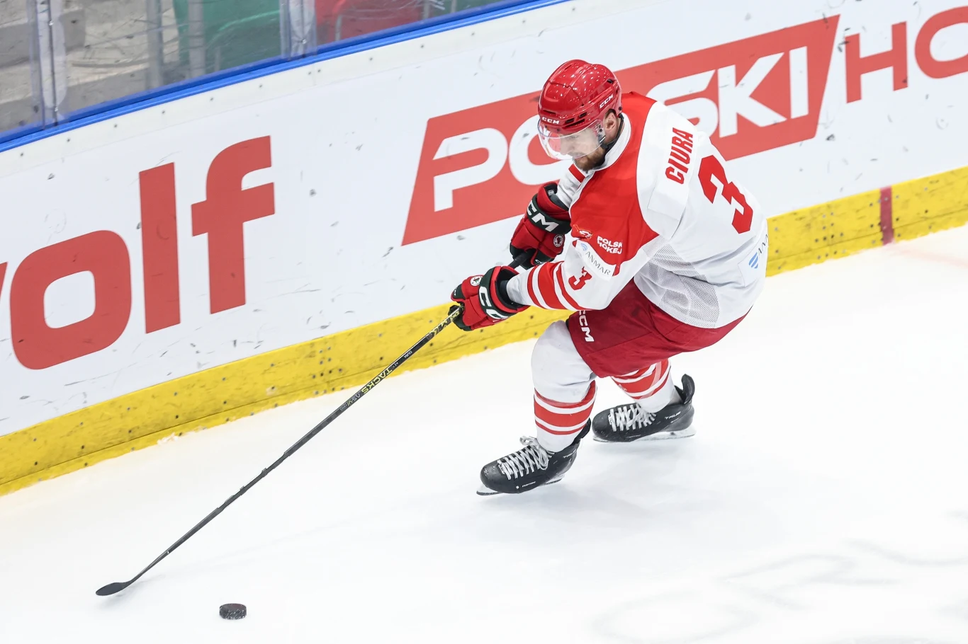 Hokeista w czerwono-białym stroju z numerem 3 na plecach, trzymający kij i przygotowujący się do odbicia krążka na lodowisku, w tle widoczna biało-czerwona reklama na bandzie.