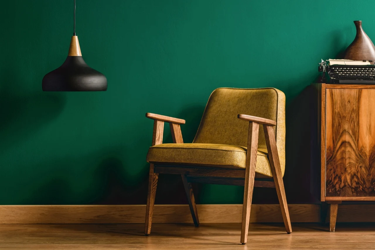 żółty fotel w stylu retro ustawiony przy drewnianej komodzie na tle zielonej ściany, nad fotelem zawieszona nowoczesna czarna lampa wisząca