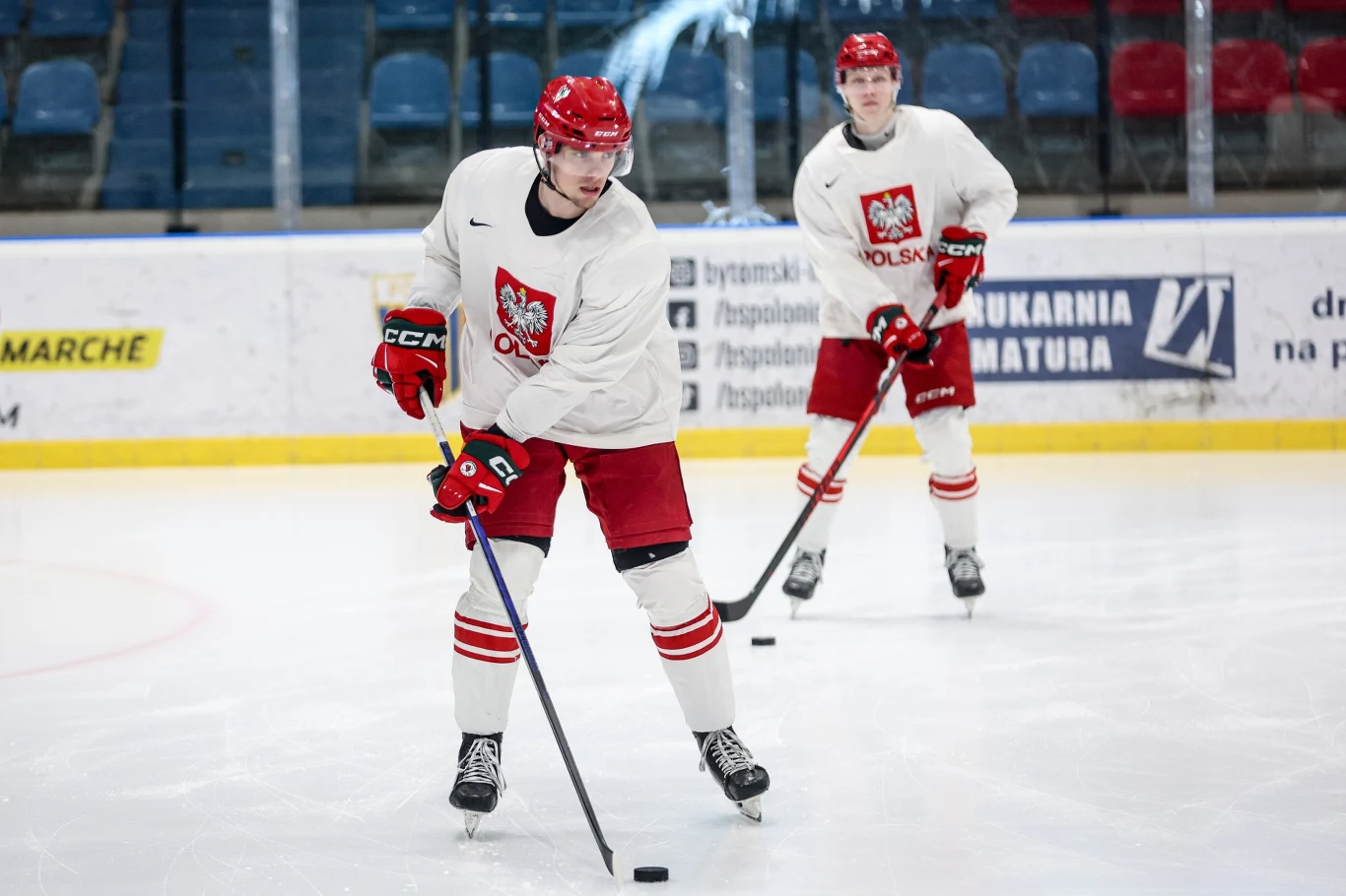 dwóch hokeistów w białych strojach z orłem na piersi oraz czerwonych spodenkach trenuje na lodowisku; zawodnik z przodu prowadzi kijem krążek, drugi obserwuje sytuację z tyłu