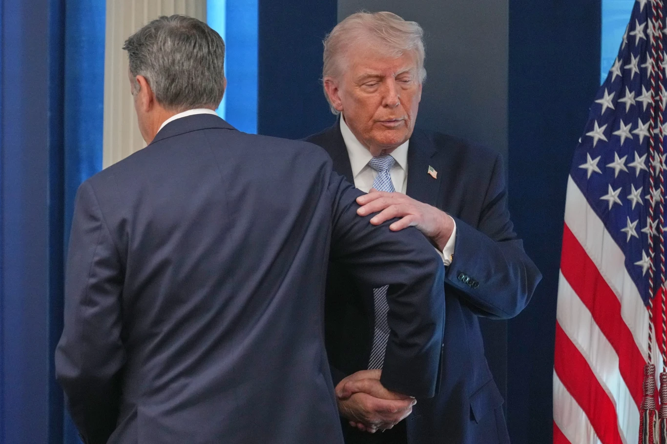 Donald Trump w garniturze dotyka łokcia mężczyzny stojącego tyłem; obok stoi flaga Stanów Zjednoczonych na tle niebieskich paneli.