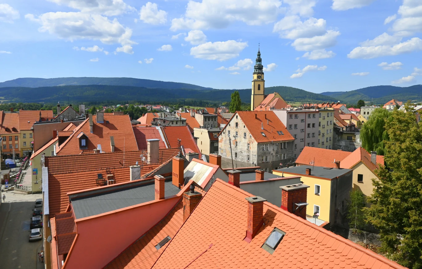 Dlaczego Bystrzyca Kłodzka nazywana jest "polską Toskanią"? Czerwone dachy kamienic w Bystrzycy Kłodzkiej, wieża kościoła z zegarem i panorama gór pod niebem z chmurami
