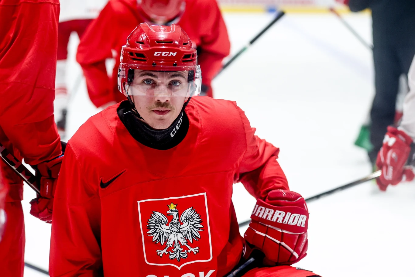 hokeista w czerwonym stroju z godłem Polski na piersi i czerwonym kasku, klęczy na lodowisku podczas treningu, otoczony innymi zawodnikami
