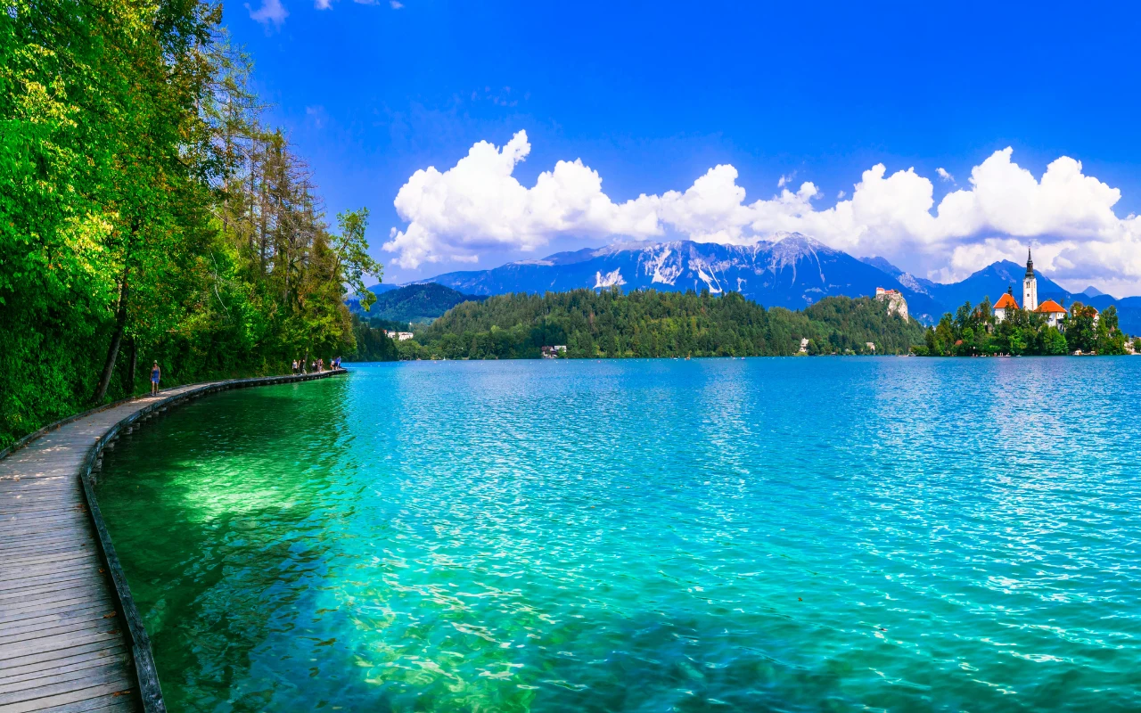 Promenada przy jeziorze Bled Turkusowe jezioro otoczone bujną zielenią, drewniana ścieżka biegnąca wzdłuż brzegu, w oddali wyłaniające się ośnieżone szczyty gór oraz kościół i wieża na małej wyspie pod białymi chmurami na tle błękitnego nieba.