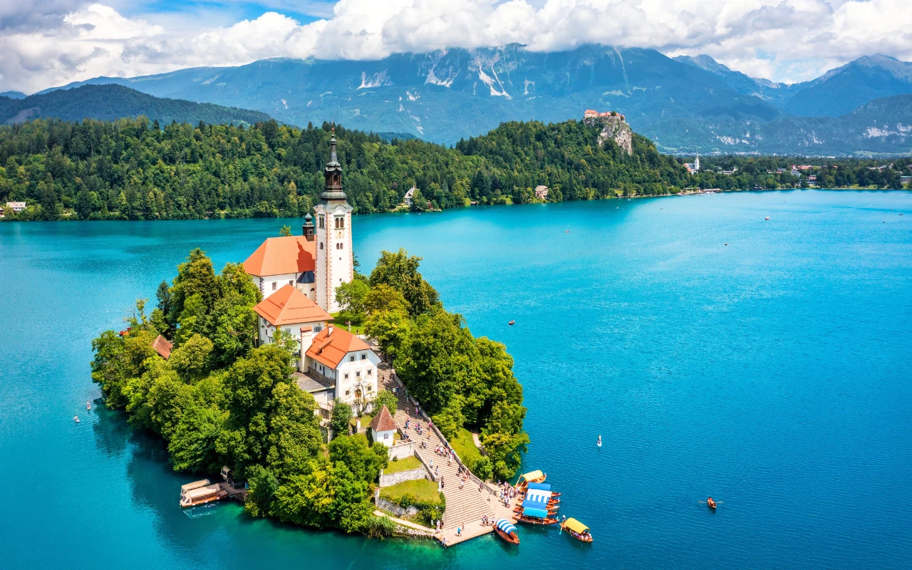 Kościół Wniebowzięcia Najświętszej Maryi Panny malownicza wyspa na środku jeziora z kościołem o charakterystycznej wieży, otoczona gęstą zielenią i turkusową wodą, w tle rozciągają się lasy, wzgórza oraz majestatyczne góry pod częściowo zachmurzonym niebem