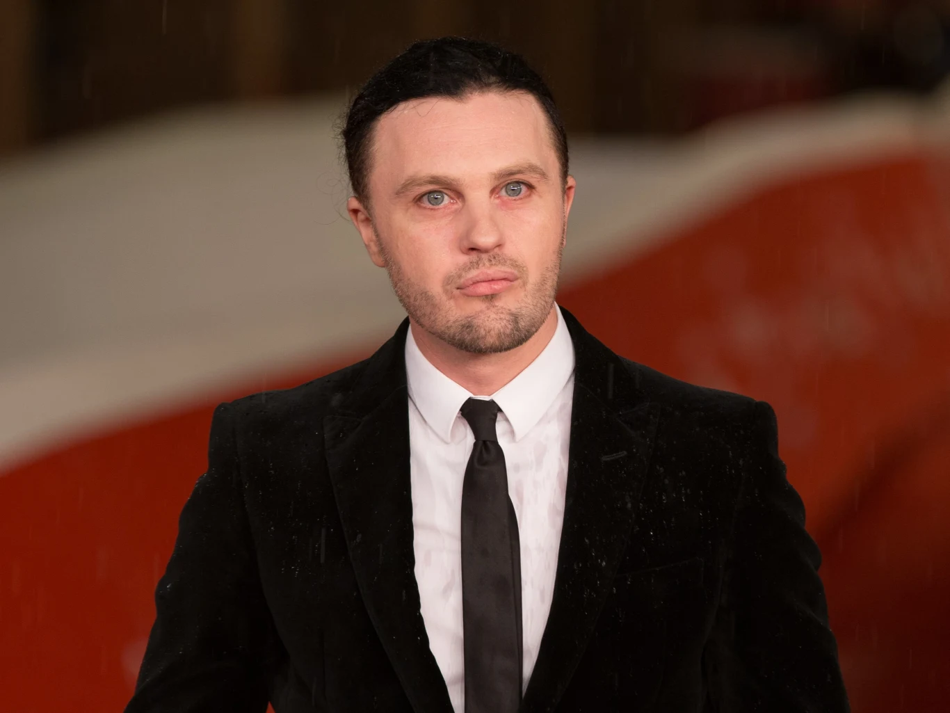 Michael Pitt Mężczyzna ubrany w elegancki garnitur stoi na tle dużej czerwono-białej formy w rozmytym tle