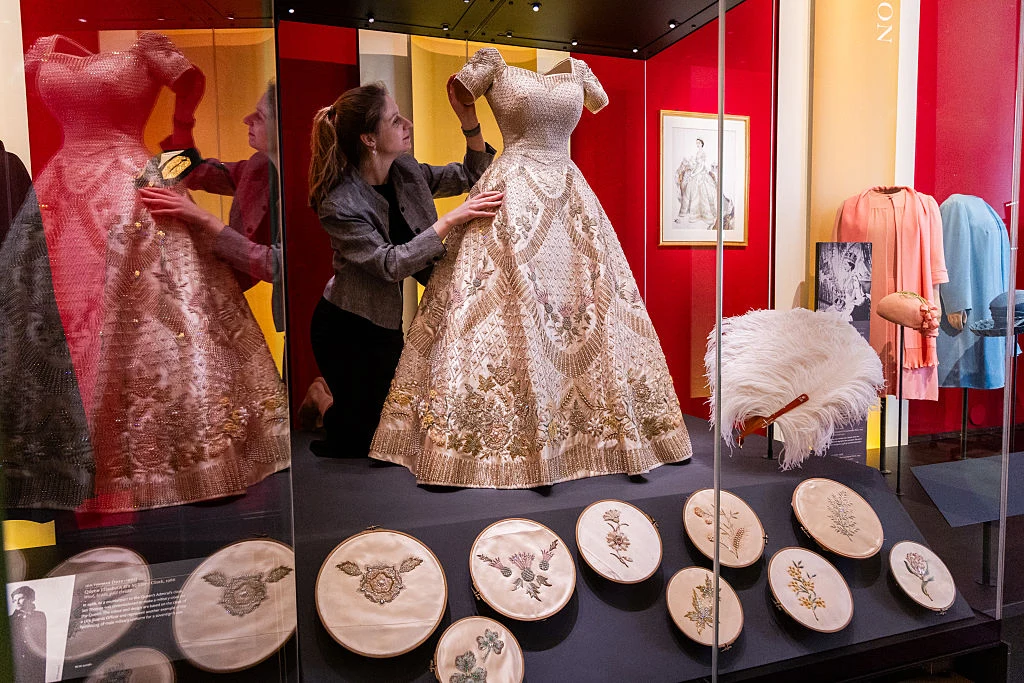 Suknia koronacyjna królowej Elżbiety II na wystawie "Queen Elizabeth II: Her Life in Style" rozłożysta, bogato zdobiona suknia balowa eksponowana w szklanej gablocie, kobieta dokładnie ją ogląda i poprawia detale, wokół rozmieszczone hafty w tamborkach, obok wielkie białe pióro oraz inne elementy garderoby na ekspozycji muzealnej
