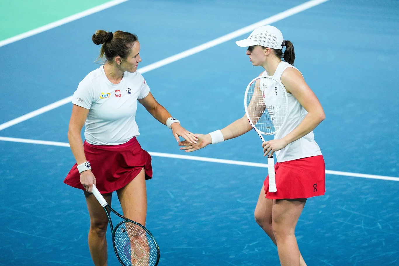 Dwie tenisistki w białych koszulkach i czerwonych spódnicach wymieniają uścisk dłoni na korcie tenisowym, trzymając rakiety i pokazując sportową współpracę.