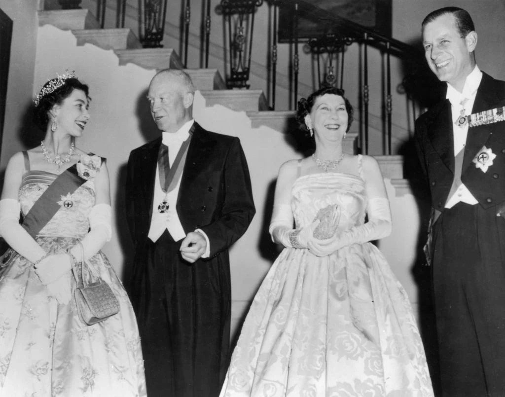 Od lewej: królowa Elżbieta II, Dwight Eisenhower, jego żona Mamie i książę Filip. Większość suknię z 1957 roku zna wyłącznie z czarno-białych zdjęć, podobnie jak wiele kreacji królowej z tego okresu Cztery uśmiechnięte osoby w eleganckich strojach wieczorowych, dwie kobiety w sukniach, tiarach i rękawiczkach oraz dwóch mężczyzn w smokingach i z odznaczeniami, stoją razem na tle schodów ozdobionych kutą balustradą.