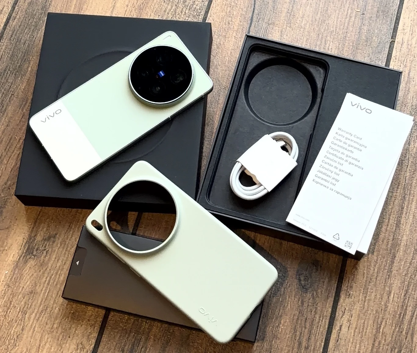 dwa smartfony Vivo z dużymi, okrągłymi modułami aparatów, biały kabel USB oraz karta informacyjna na tle otwartych czarnych pudełek, wszystko ułożone na drewnianej powierzchni