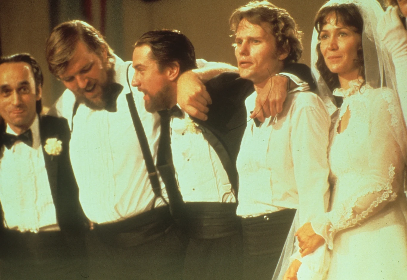 Kadr z filmu "Łowca jeleni". Pierwszy z lewej to John Cazale Grupa mężczyzn w smokingach z ramionami opartymi na sobie, obok nich kobieta w welonie ślubnym i białej sukni, wszyscy wyglądają na wzruszonych i zjednoczonych, scena sugeruje ważne rodzinne lub przyjacielskie wydarzenie.