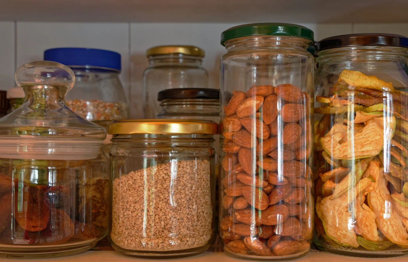 Szklane słoiki ustawione na półce, wypełnione suszonymi owocami, migdałami oraz sezamem, każdy z nich zamknięty metalową lub plastikową zakrętką.