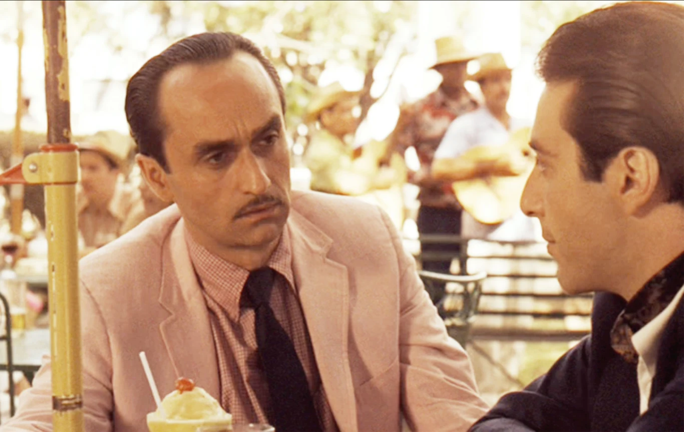 John Cazale jako Fredo Corleone i Al Pacino jako Don Michael Corleone w filmie "Ojciec Chrzestny II" (1974) Dwaj mężczyźni w garniturach siedzą naprzeciwko siebie przy stoliku na świeżym powietrzu, jeden z nich ma poważny wyraz twarzy i pozuje w jasnoróżowej marynarce oraz krawacie, na stole stoi napój z cytryną i słomką, w tle widać muzyków w sombrero oraz ...