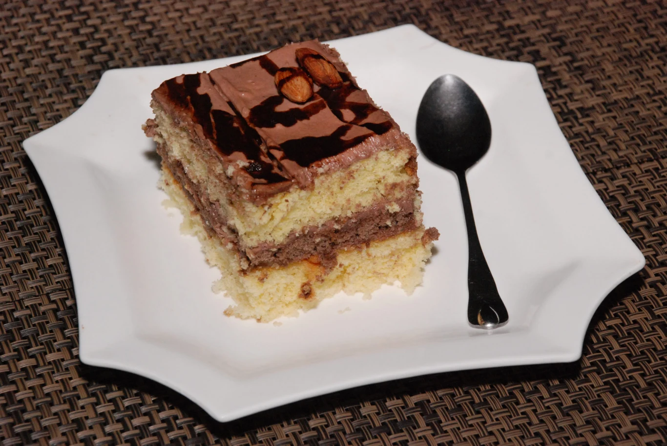 Czekoladowe ciasto z dodatkiem śliwek na rodzinne imprezy Kawałek warstwowego ciasta czekoladowo-śliwkowego z polewą i orzechami na białym talerzu, obok czarna łyżeczka.