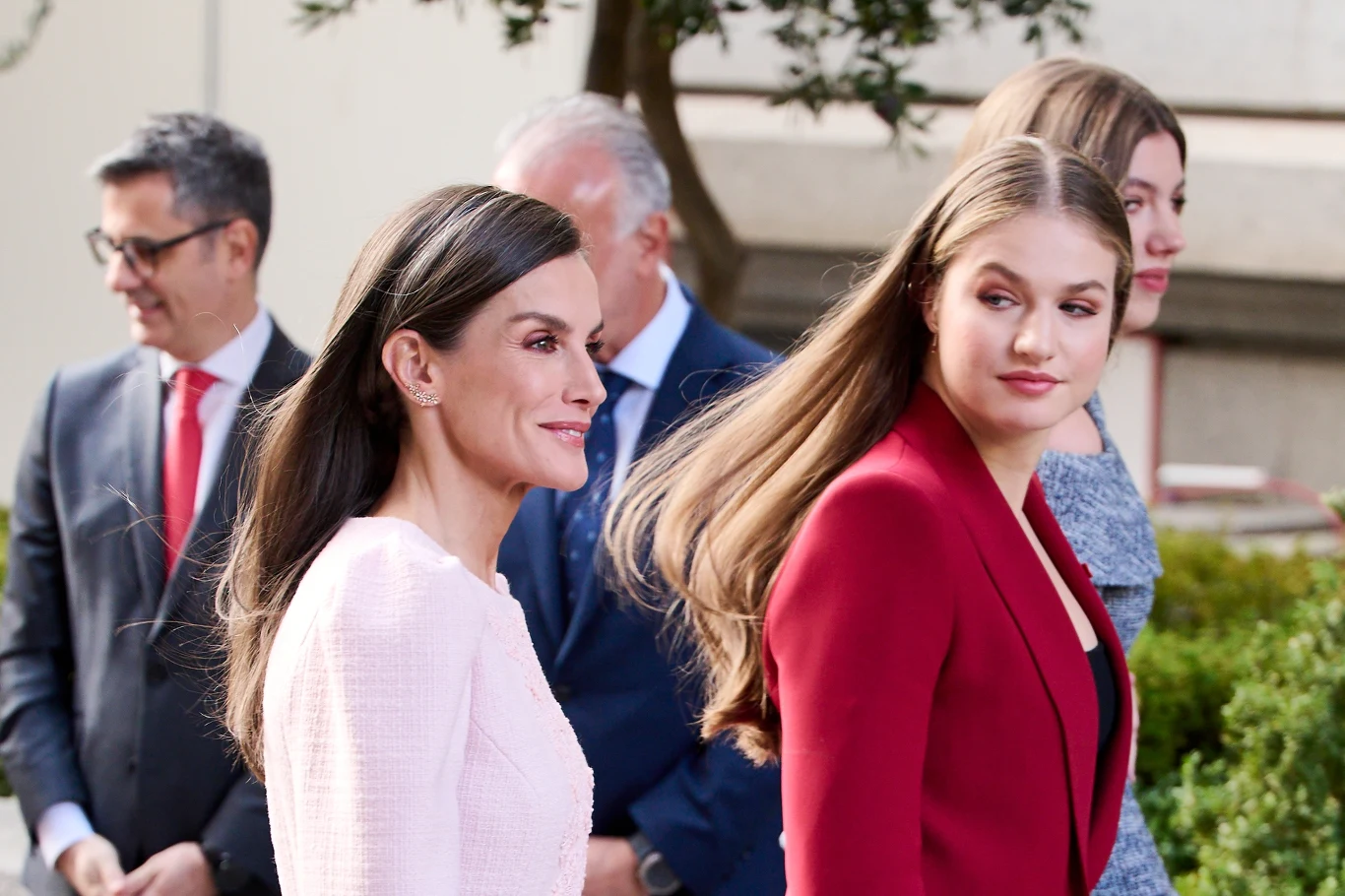 Młodsza córka często szuka wzrokiem matki, a ta wygląda jakby ją ignorowała. Czy to przypadek? Dwie elegancko ubrane kobiety idą obok siebie, jedna w jasnoróżowym żakiecie, druga w bordowej marynarce, na drugim planie kilku mężczyzn w garniturach i kolejna kobieta, wokół widoczna zieleń ogrodowa i budynek w tle.