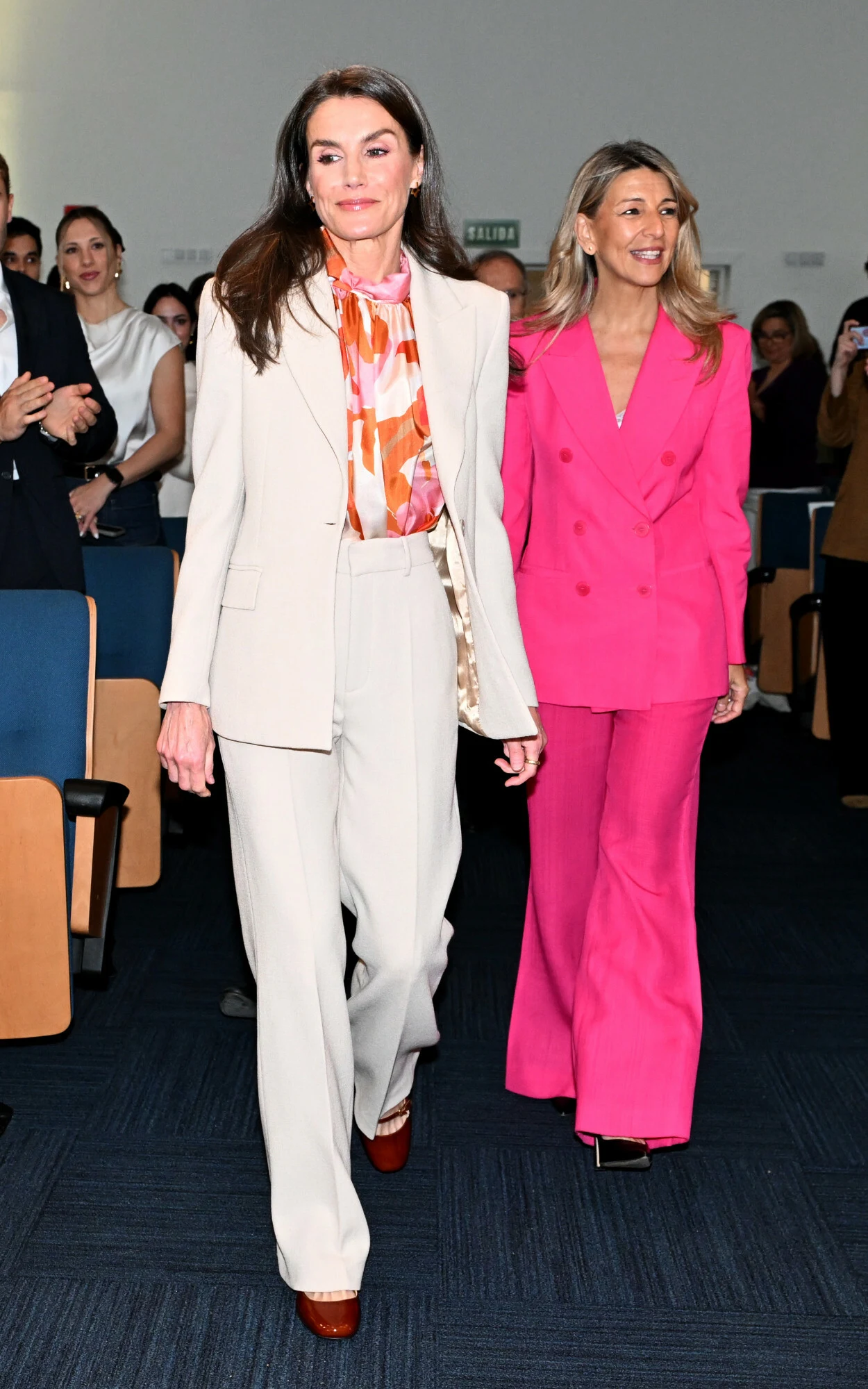 Dwie elegancko ubrane kobiety idą alejką wśród siedzących ludzi, pierwsza w jasnym garniturze i kolorowej koszuli, druga w intensywnie różowym garniturze; obie z pewnością siebie, otoczone przez klaszczącą publiczność.