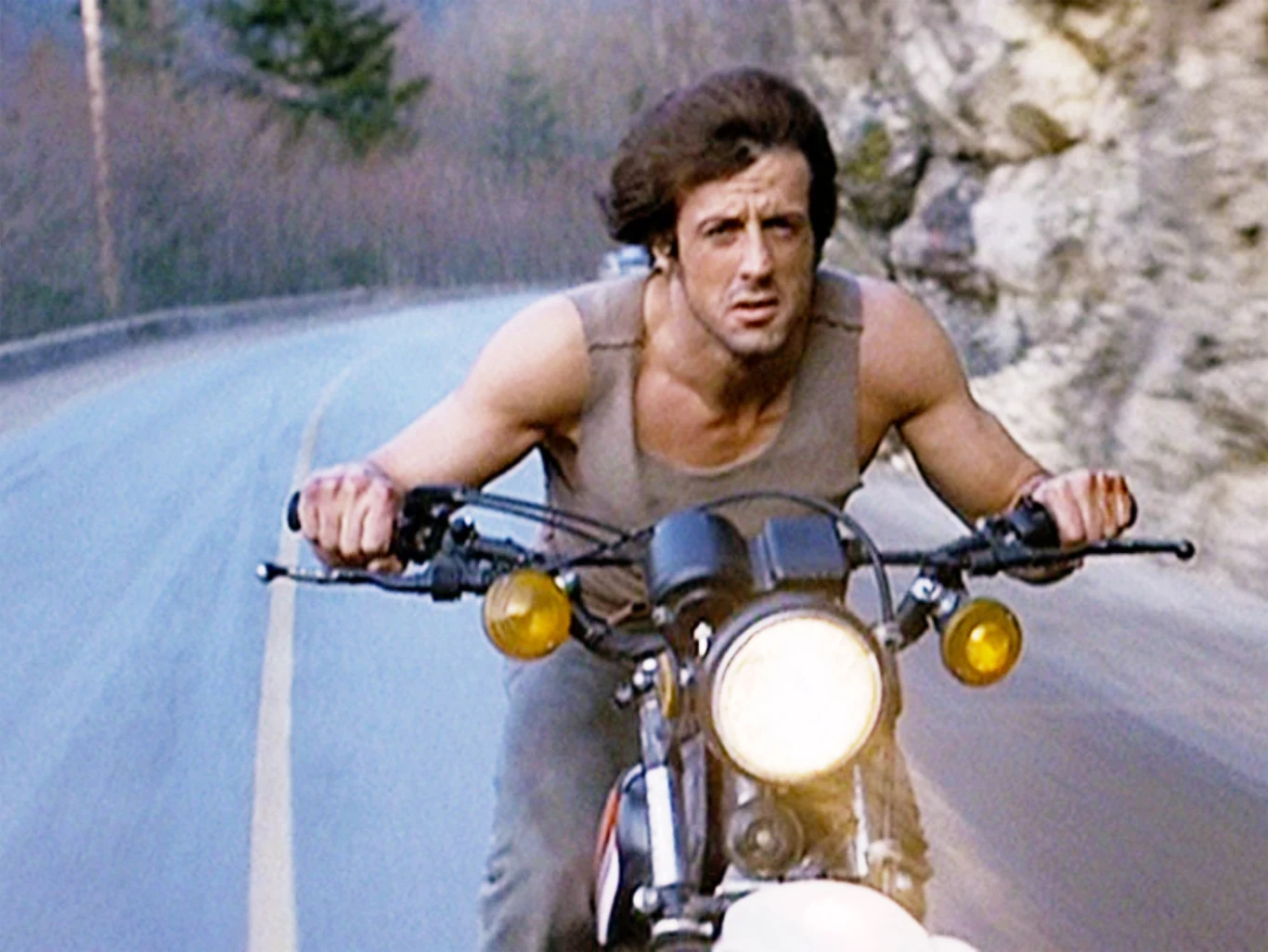 Sylvester Stallone w filmie "Rambo: Pierwsza krew" (1982) mężczyzna o muskularnej sylwetce w szarej koszulce jedzie motocyklem po wąskiej, krętej drodze otoczonej drzewami i skałami, patrząc intensywnie przed siebie