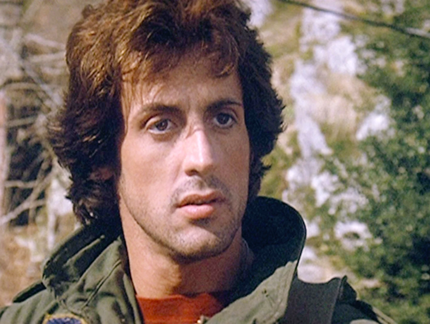 Sylvester Stallone w filmie "Rambo: Pierwsza krew" (1982) Mężczyzna o ciemnych, kręconych włosach w zielonej kurtce patrzy przed siebie z poważnym wyrazem twarzy, w tle rozmyte drzewa i skały.